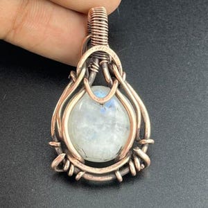 May include: A teardrop-shaped pendant featuring a smooth, translucent white moonstone cabochon encased in intricate, hand-wrapped copper wire. The wire forms a decorative frame around the stone with a coiled bail at the top.