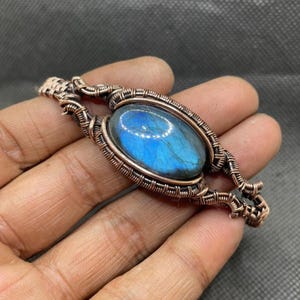 May include: A wire-wrapped bracelet featuring a central oval labradorite gemstone with a vibrant blue iridescent flash. The stone is encased in intricate, hand-woven copper wire detailing that extends along the band, creating a textured metal frame.