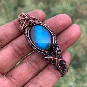 May include: A bracelet featuring a central oval labradorite gemstone with a vibrant blue flash, encased in an intricate, hand-woven copper wire frame. The metalwork consists of multiple coiled and twisted copper strands surrounding the stone.