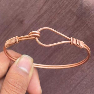 May include: A copper wire bracelet featuring a double-strand design that forms a teardrop-shaped hook closure. The wire is wrapped tightly around the band at two points, creating a secure connection for the hook and eye mechanism.