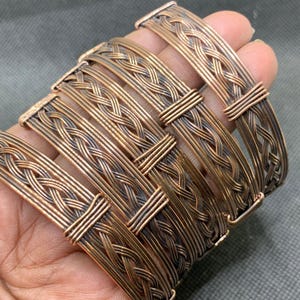 Könnte beinhalten: Ein Set aus vier kupferfarbenen Metallarmbändern mit geflochtenem Drahtmuster. Jedes Armband hat ein flaches, breites Banddesign mit geflochtener Textur und ist an den Enden mit dünnen Drahtwicklungen befestigt, mit einem dunkel oxidierten Finish.