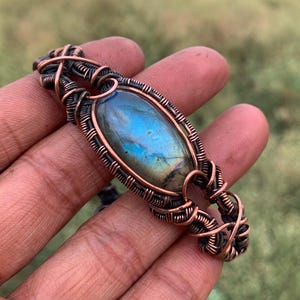 May include: A bracelet featuring an oval labradorite gemstone with blue iridescence, encased in a complex, dark-toned copper wire-wrapped setting. The metalwork consists of multiple intertwined strands of wire forming a decorative, cage-like structure.