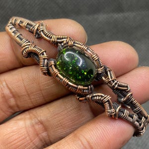 May include: A handcrafted copper wire bracelet featuring a central oval-shaped green stone with sparkling inclusions. The wire is intricately woven, creating a unique, textured design. The bracelet is a piece of jewelry.