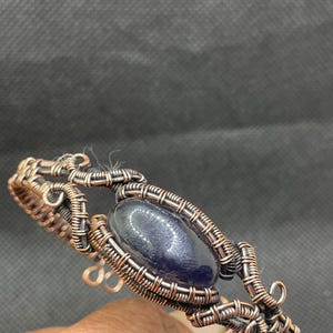 May include: A handcrafted copper wire bracelet featuring a dark blue oval stone at the center. The wire is intricately woven around the stone and forms a decorative openwork design. The bracelet is a unique piece of jewelry.