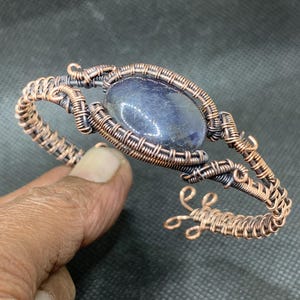 May include: A handcrafted copper wire bracelet featuring a central oval-shaped, dark blue gemstone. The wire is intricately woven and coiled, creating a textured, open design. The bracelet has an adjustable opening.