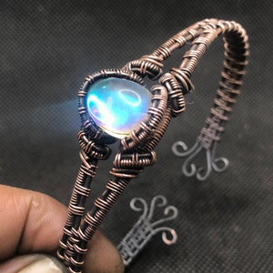 May include: A copper wire wrapped bracelet with a iridescent blue and green gemstone. The bracelet is made with a woven wire design.
