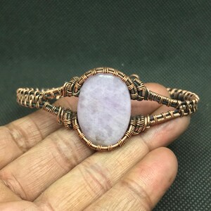 May include: A copper wire wrapped bracelet featuring a light purple oval gemstone in the centre.