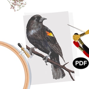 May include: A cross-stitch pattern of a black bird with a yellow and orange wing patch perched on a branch. The image includes embroidery floss in yellow, red, and black, a needle, and a wooden embroidery hoop. A PDF icon is visible.