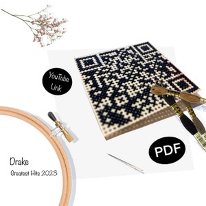 May include: Cross stitch pattern of a black and white QR code with the text "Drake Greatest Hits 2023" and "PDF" and "YouTube Link". The pattern is on a white background with a wooden embroidery hoop, embroidery floss, and a needle.