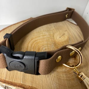Op de afbeelding: Een bruine hondenriem met een zwarte gesp en goudkleurige hardware. De halsband heeft een getextureerd oppervlak en een kleine, crèmekleurige kwast. De halsband is ontworpen voor huisdieren.