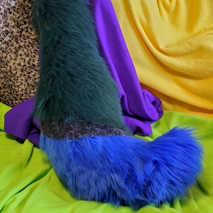 Può includere: Un accessorio a coda in peluche realizzato in pelliccia sintetica lunga, caratterizzato da una sezione superiore verde bosco scuro, una stretta fascia grigio antracite al centro e una sezione inferiore blu reale vivace che si curva verso l'esterno.