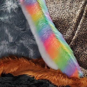 May include: A long, tapered tail accessory made of soft, multi-coloured faux fur featuring a vibrant rainbow gradient of pink, orange, yellow, green, blue, and purple hues. The plush, textured surface mimics animal fur with a dense, fluffy pile.