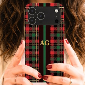 May include: A smartphone case featuring a red, green, and black plaid pattern with a vertical dark brown stripe down the center. The letters AG are printed in gold in the middle of the stripe. The case has a cutout for a triple camera system.