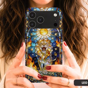 May include: A smartphone case featuring a stained glass style design of a roaring lion centered beneath a bright sun. The artwork incorporates deep blue, gold, and orange tones with star motifs and flame-like patterns. The word LEO is printed in a small black box.