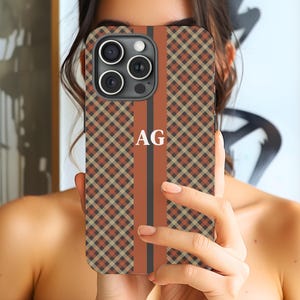 May include: A smartphone case featuring a diagonal plaid pattern in shades of brown, tan, and black. A vertical solid rust-colored stripe runs down the center, containing the white capital letters AG. The case has a cutout for a triple-lens camera.
