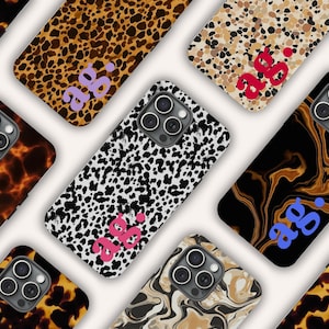 May include: A collection of smartphone cases featuring various patterns including leopard print, tortoiseshell, and marble designs. Each case has a triple-lens camera cutout and the lowercase letters ag. printed in contrasting colours on the back.