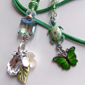 May include: Two green beaded bookmarks with charms. One has a large iridescent square bead and a cluster of crystals, a white flower, and a gold leaf. The other has a green turtle charm and a green butterfly charm. The bookmarks have green cords.