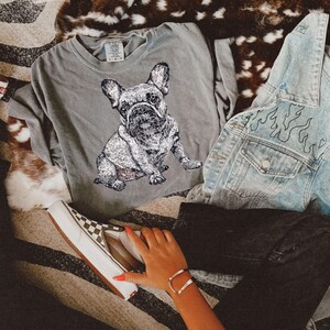 May include: A gray t-shirt with a black and white image of a French bulldog. The shirt is laying on a brown and white patterned surface. A denim jacket with a black flame design is laying on top of the shirt. A person is wearing black pants and a pair of checkered Vans shoes.