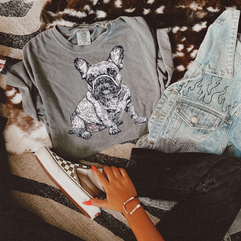 May include: A gray t-shirt with a black and white image of a French bulldog. The shirt is laying on a brown and white patterned surface. A denim jacket with a black flame design is laying on top of the shirt. A person is wearing black pants and a pair of checkered Vans shoes.