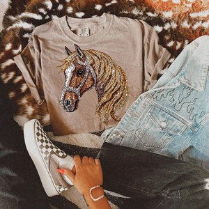May include: A light brown t-shirt with a rhinestone horse design. The horse is brown and gold with a brown bridle. The shirt is worn with a denim jacket and black pants.