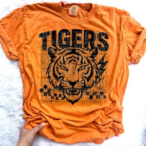 May include: Orange Comfort Colors t-shirt with a black tiger graphic and the word "TIGERS" above it. The tiger graphic has a distressed design. The shirt has long sleeves and a crew neck.