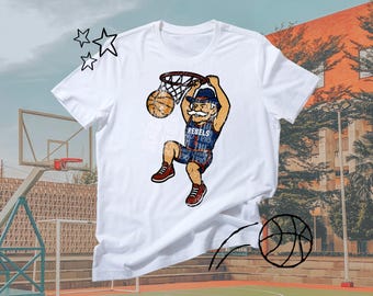 Puede incluir: Camiseta blanca con un jugador de baloncesto de dibujos animados haciendo un mate. El jugador lleva un uniforme azul y rojo con la palabra "REBELS". El balón de baloncesto es naranja y marrón. La camiseta tiene texto.