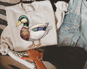 May include: A white sweatshirt with a rhinestone-encrusted duck design. The duck is green, brown, and white with a purple and green iridescent sheen. The sweatshirt is on a denim jacket and a pair of black pants.