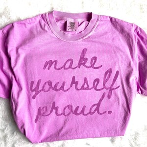 Puede incluir: Una camiseta Comfort Colors de color lila claro con la frase "make yourself proud" en una escritura morada m&aacute;s oscura. La camiseta tiene cuello redondo y mangas cortas. La etiqueta dice "L-G".