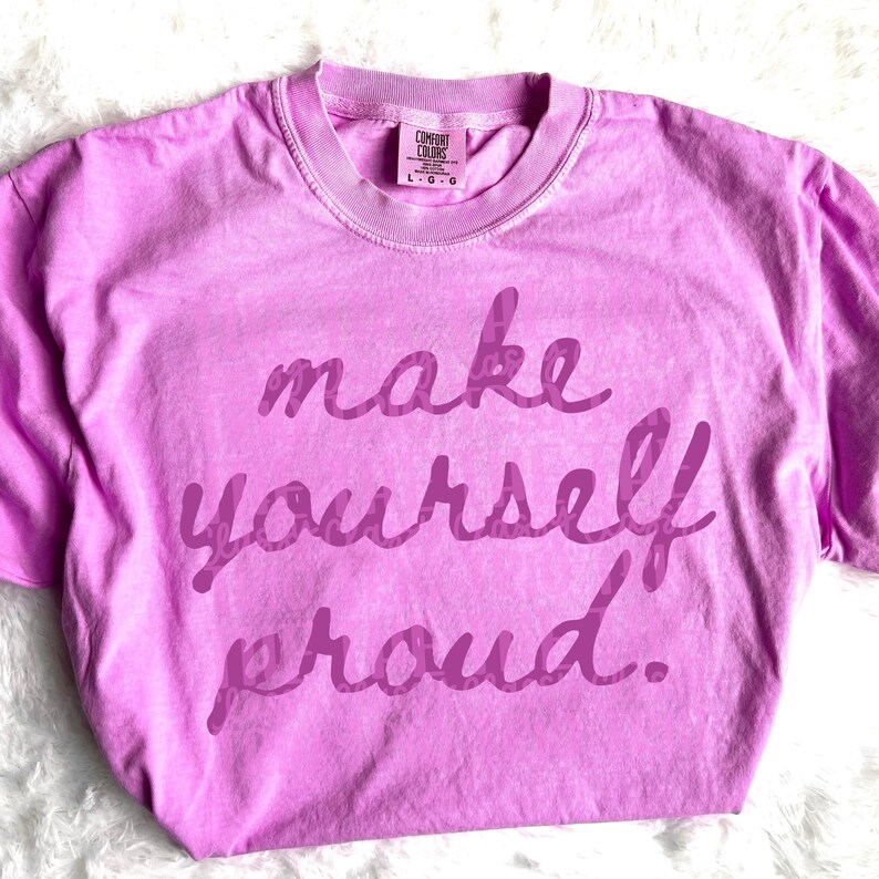 Puede incluir: Una camiseta Comfort Colors de color lila claro con la frase "make yourself proud" en una escritura morada m&aacute;s oscura. La camiseta tiene cuello redondo y mangas cortas. La etiqueta dice "L-G".