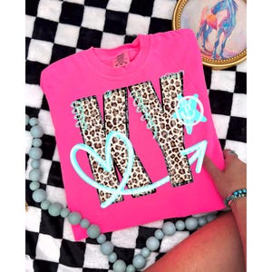 May include: A bright pink sweatshirt featuring the word "KIND" in a leopard print pattern. The word is outlined in light blue, with a heart and arrow design. The shirt is displayed on a black and white checkered surface.