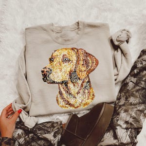 May include: A beige sweatshirt with a brown and gold sequined dog design. The dog is a Labrador Retriever with its head turned to the side. The sweatshirt is laying on a brown and white patterned fabric.