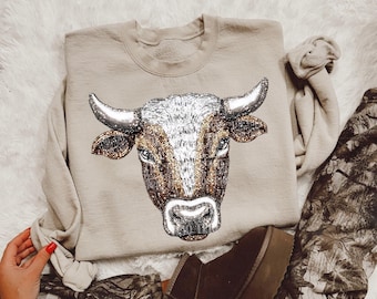 Puede incluir: Una sudadera beige con un diseño detallado de cabeza de toro. La cabeza del toro está representada en tonos marrones, blancos y dorados, con un aspecto texturizado. La sudadera se combina con pantalones de camuflaje y botas marrones.