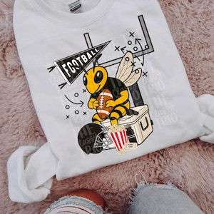 May include: White sweatshirt with a cartoon bee holding a football, set against a football-themed backdrop. The design includes a helmet, popcorn, and the words "FOOTBALL" and "THE CLASSY TRIO".
