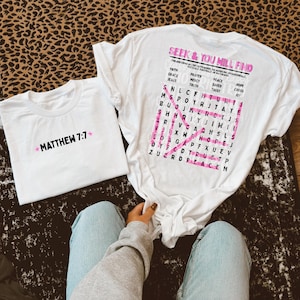 May include: Two white t-shirts. One features the black text "MATTHEW 7:7" with pink stars. The other displays a pink word search puzzle titled "SEEK & YOU WILL FIND" with words like "FAITH," "PRAYER," "PEACE," "GRACE," "MERCY," "SAVED," "JESUS," "TRUTH," "TRUST," "HOPE," "CROSS," and "JOY" printed on the back.
