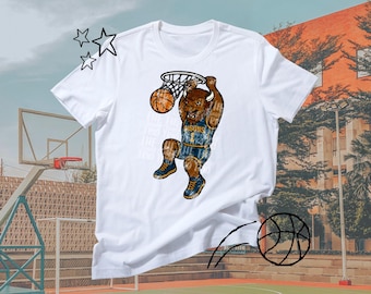 Puede incluir: Camiseta blanca con un bisonte de dibujos animados encestando una pelota de baloncesto. El bisonte lleva una camiseta azul y dorada con la palabra "Bisons". La camiseta tiene texto en la parte delantera y está sobre un fondo de cancha de baloncesto.