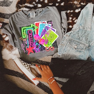 May include: A gray t-shirt with a neon graphic design featuring a horse, playing cards, cherries, and a neon sign that says "OPEN".