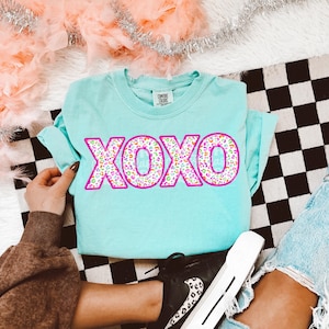 May include: A light blue t-shirt with the word "XOXO" in a pink outline and a colorful leopard print pattern. The shirt is folded, and the sleeves are rolled up. The shirt is on a black and white checkered surface.