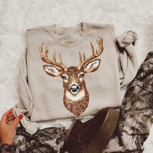 May include: A beige sweatshirt with a detailed deer head design in brown and gold. The deer has large antlers and is centered on the front. The image also shows brown boots and camouflage trousers.