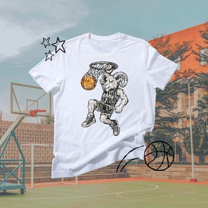 Pode incluir: Camiseta branca com uma ilustração em preto e branco de um carneiro jogando basquete. O carneiro está vestindo uma camisa com o texto "RAMS" e está enterrando uma bola de basquete. O design tem um estilo vintage. O fundo mostra uma quadra de basquete.