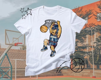 Puede incluir: Camiseta blanca con un gráfico de un jugador de baloncesto haciendo un mate. El jugador lleva un uniforme azul y rojo con la palabra "Spartans". También se muestra una red y un balón de baloncesto. El fondo representa una cancha de baloncesto.
