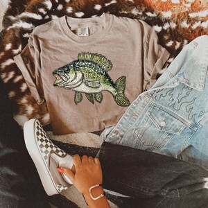 May include: A brown t-shirt with a green and black fish graphic. The fish is a bass with a large mouth and fins. The t-shirt is worn by a person wearing a denim jacket and checkered Vans shoes.