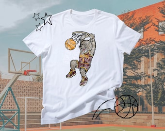 Puede incluir: Camiseta blanca con un lobo de dibujos animados haciendo un mate de baloncesto. El lobo lleva una camiseta con la palabra "WOLVES" y el número 1. Se ven una red y un balón de baloncesto. El fondo incluye una cancha de baloncesto y un edificio.