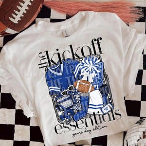 May include: A white t-shirt featuring a graphic print with the text "the kickoff essentials game day edition". The design includes a blue sweater, a pom-pom, a football, sunglasses, a cup, a towel, and a ticket stub inside a wire basket.