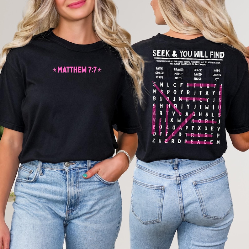 May include: A black short-sleeve t-shirt featuring pink text on the front that reads MATTHEW 7:7. The back displays a word search puzzle with the title SEEK & YOU WILL FIND in white, containing words like FAITH, GRACE, and MERCY highlighted in pink.