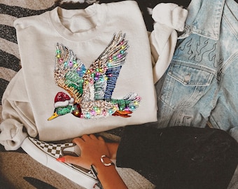 May include: A cream-colored sweatshirt featuring a detailed, bejeweled duck design with colorful wings and a Santa hat. The duck is embellished with various colors of rhinestones. A light blue denim jacket with flame designs is visible.