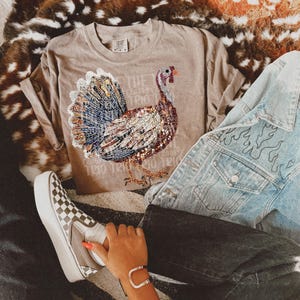 May include: A brown t-shirt with a sparkly turkey design and the text "The Turkey Whisperer" printed on it. The shirt is being worn with a denim jacket and checkered Vans shoes.