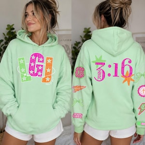 Puede incluir: Una sudadera con capucha de color verde claro que presenta un gráfico frontal con las letras LGJ en rosa, verde y naranja con estrellas. La parte posterior muestra el texto John 3:16 en rosa, rodeado de varios iconos religiosos coloridos.