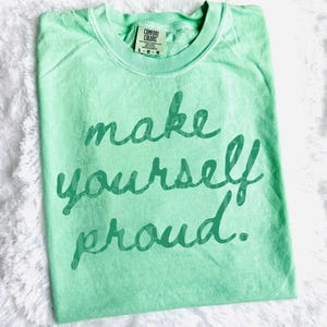 Puede incluir: Camiseta verde claro con la frase "make yourself proud" en escritura verde oscuro. La camiseta tiene cuello redondo y una etiqueta que dice "Comfort Colors". La camiseta est&aacute; sobre una superficie blanca y esponjosa.