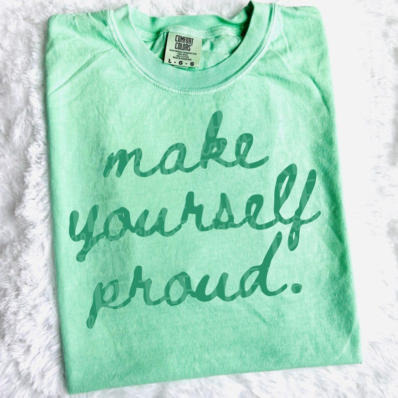 Puede incluir: Camiseta verde claro con la frase "make yourself proud" en escritura verde oscuro. La camiseta tiene cuello redondo y una etiqueta que dice "Comfort Colors". La camiseta est&aacute; sobre una superficie blanca y esponjosa.