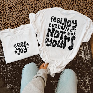 May include: Two white t-shirts with black text. One shirt says "Feel Joy" with star accents. The other says "Feel Joy even if it's NOT yours" with star accents. The shirts are on a patterned surface.